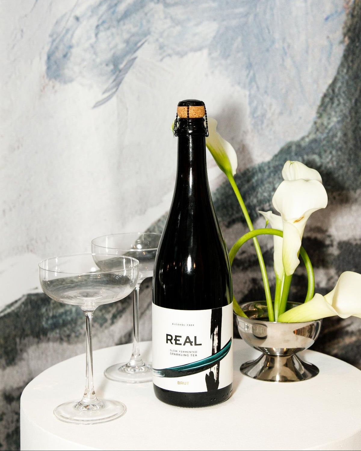 A bottle of REAL Brut alcohol-free sparkling tea on a white table with two coupes and white calla lilies