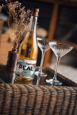 A bottle of REAL Mei sparkling tea with a cork stopper, set on a woven tray next to two coupe glasses and a small vase of dried flowers.