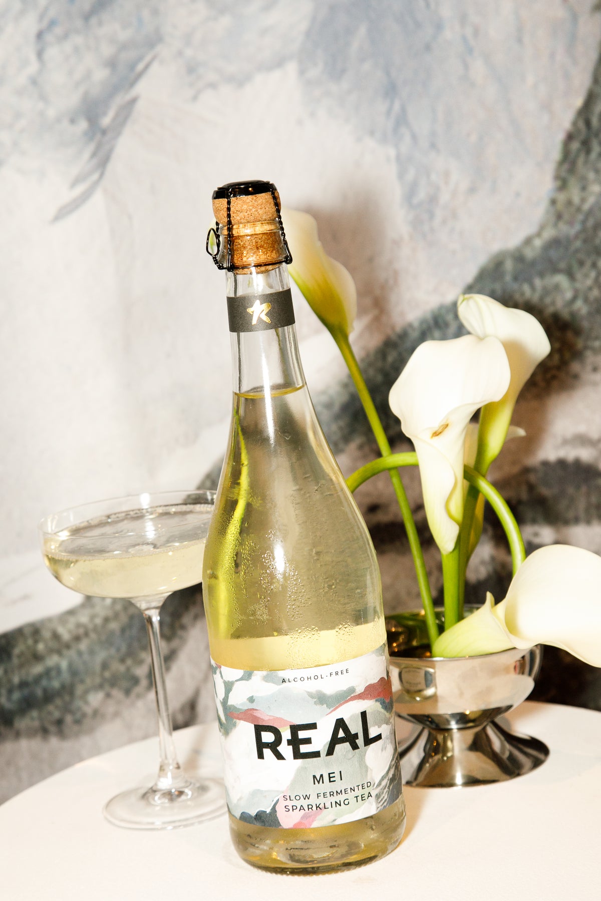 A bottle of REAL Mei sparkling tea with a gold and white label, styled on a white table next to a coupe glass filled with the tea and white calla lilies in a vase.