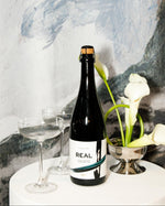 A bottle of REAL Brut alcohol-free sparkling tea on a white table with two coupes and white calla lilies