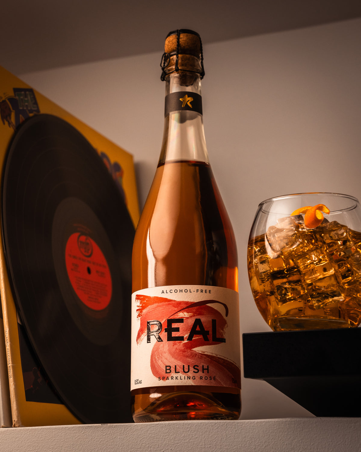 Bottle of 'Real Blush' sparkling rose with a glass of cocktail and vinyl record in the background.