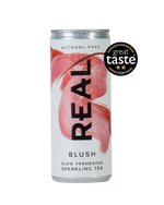 A silver aluminum can of REAL Alcohol-Free Blush Slow Fermented Sparkling Tea, featuring a pink brushstroke design and a black 'REAL' logo. A black 'great taste 2026' award badge is displayed on the top right of the can against a solid white background.