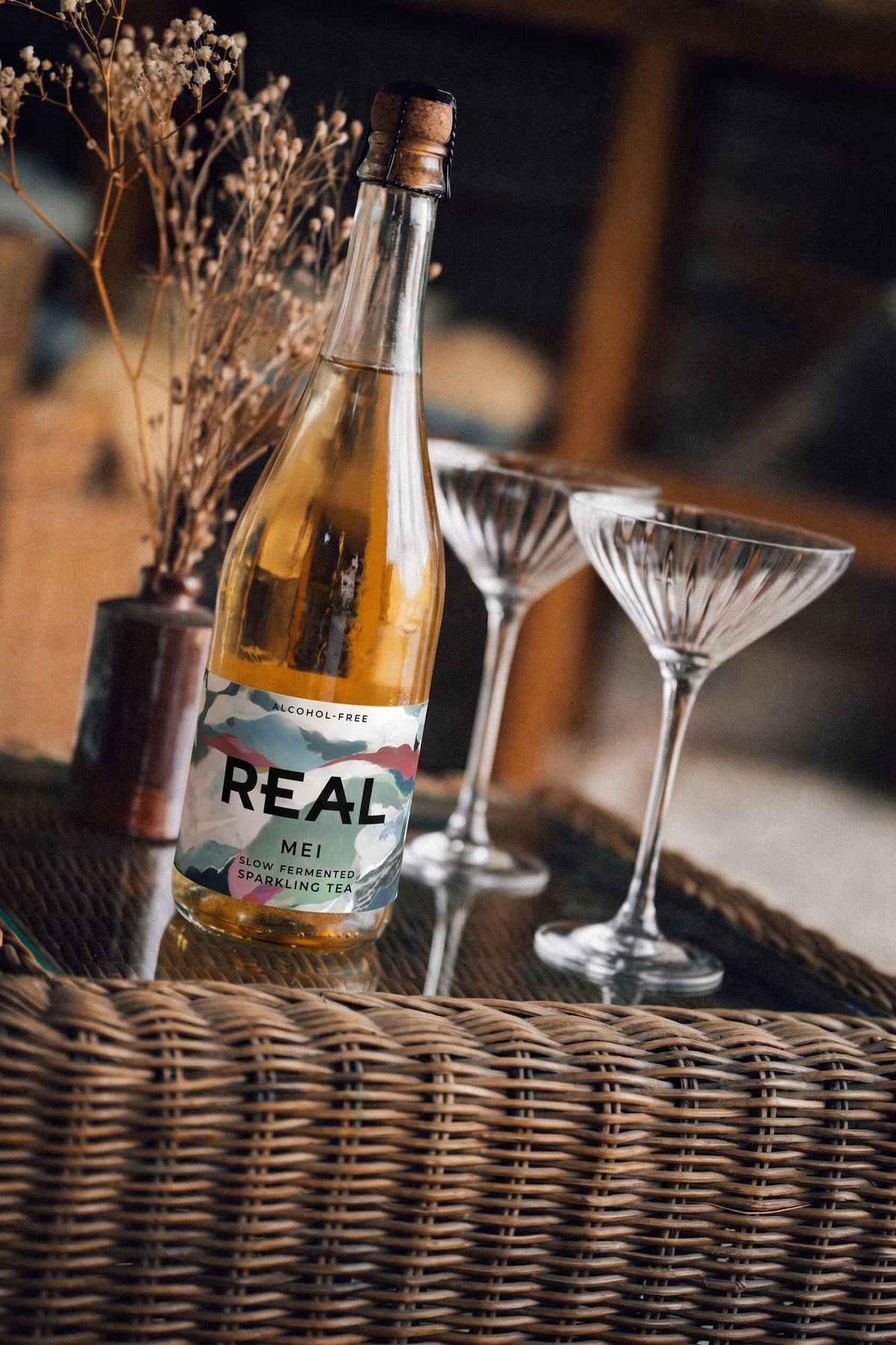 A bottle of REAL Mei sparkling tea with a cork stopper, set on a woven tray next to two coupe glasses and a small vase of dried flowers.