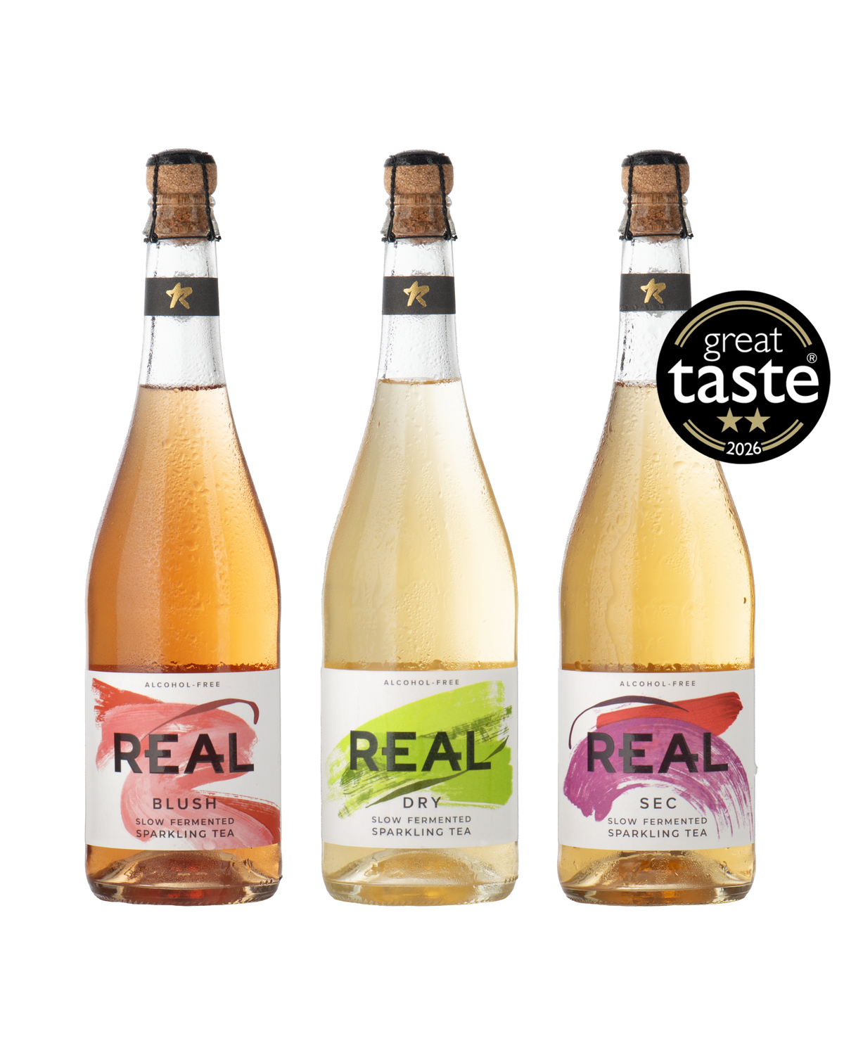 Three bottles of REAL brand sparkling tea arranged side by side on a white background, with a black 'great taste 2026' award seal overlay on the right bottle. The bottles contain pale pink, yellow, and golden-amber liquids and have white labels with red, green, and purple brushstroke accents.